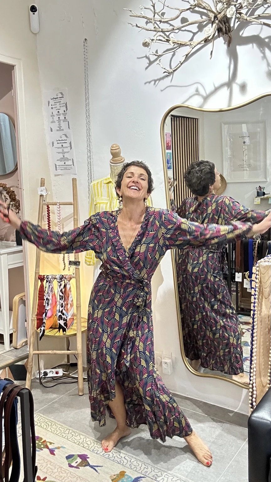 Céline souriante en robe imprimée, les bras ouverts dans sa boutique